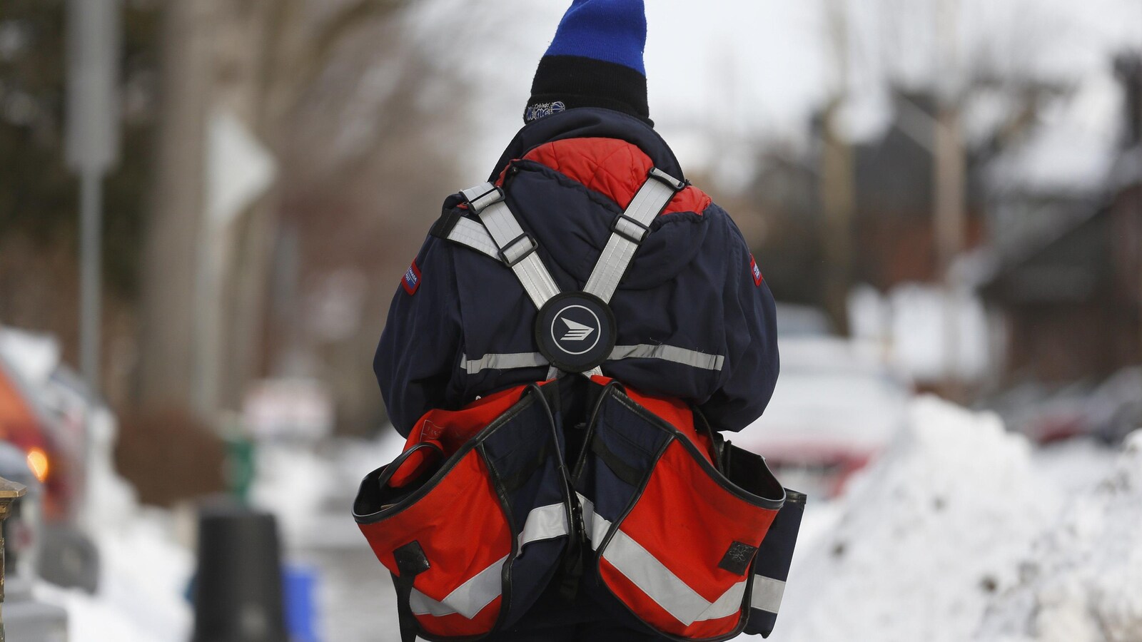 Un facteur vu du dos marche sur un trottoir enneigé.