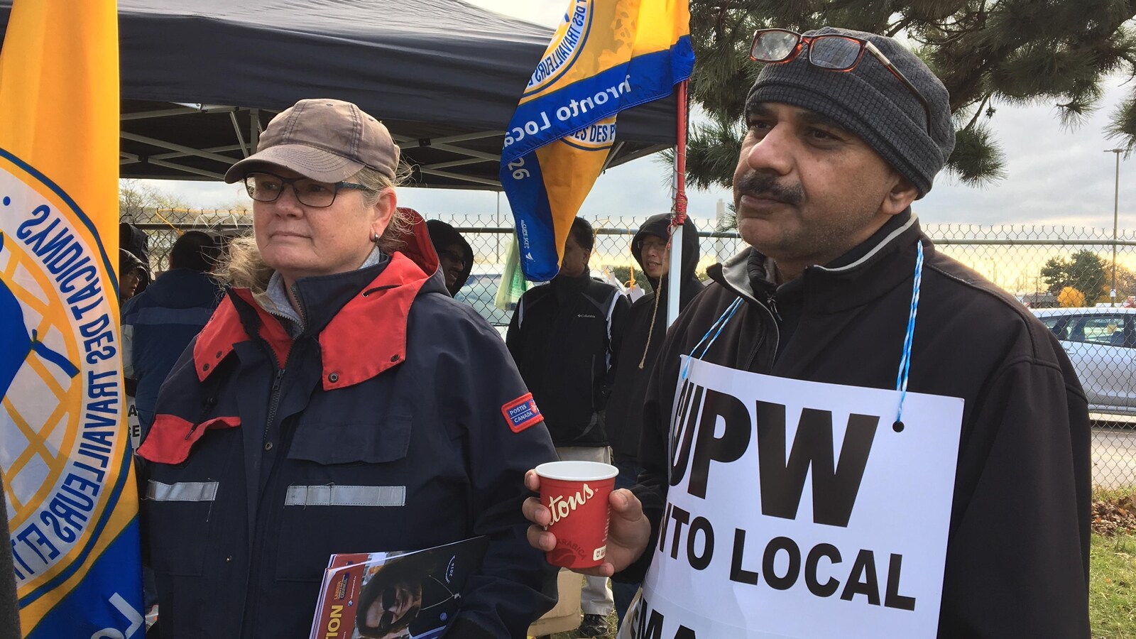 Photo d'une femme qui porte l'uniforme bleu et rouge de Postes Canada et un homme portant une affiche de son syndicat.