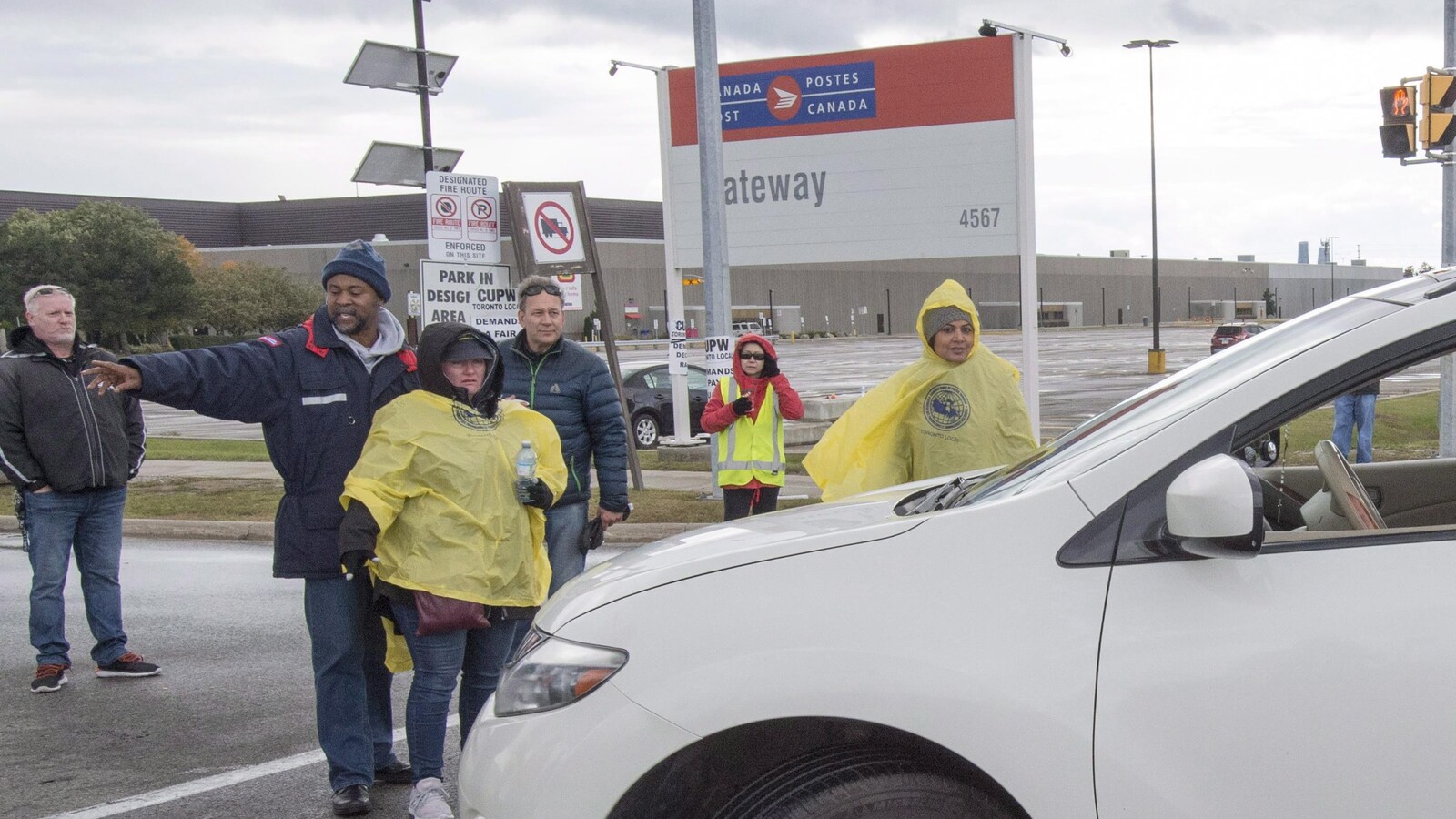 Trois femmes et deux hommes forment une ligne pour bloquer un véhicule devant un centre de tri de Postes Canada.
