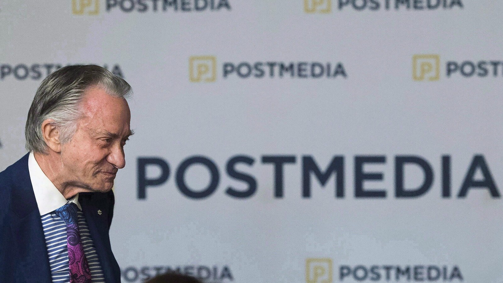 Un homme, vu de profil, se tient devant une affiche sur laquelle il est écrit « Postmedia » en lettres majuscules.