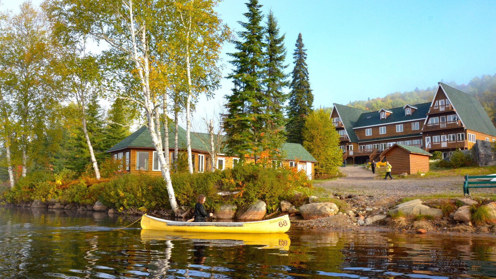 Une femme en canot sur un lac. Un grand chalet de bois et une montagne en arrière-plan.