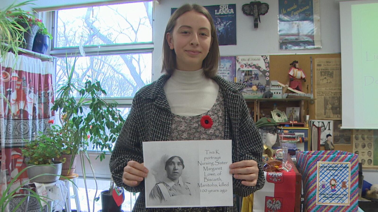Dans une classe, une adolescente aux cheveux bruns mi-longs tient une feuille sur laquelle figure le dessin d'une infirmière de l'époque, annoté du texte suivant : Tiva K portrays Nursing Sister Margaret Lowe, of Biscarth, Manitoba, killed 100 years ago.