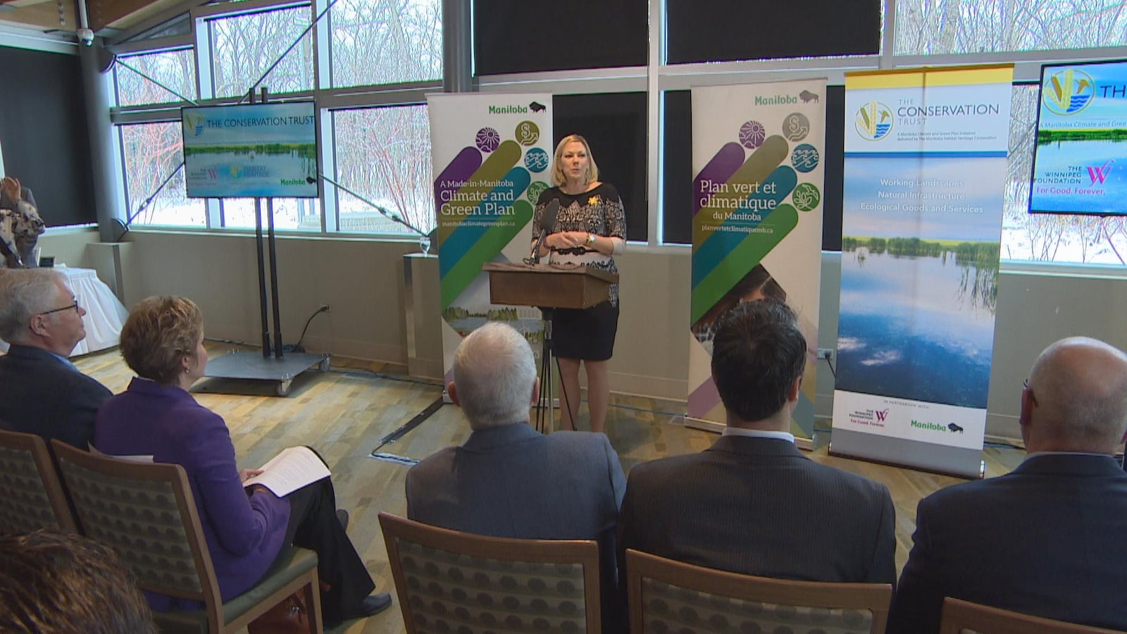 La ministre Rochelle Squires s'adresse à une foule derrière un lutrin et devant des panneaux annonçant le plan vert et climatique du Manitoba.