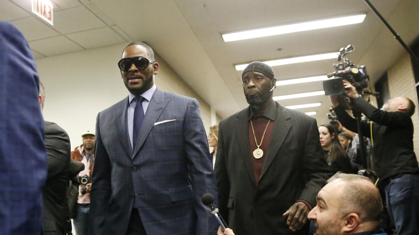 Le chanteur R. Kelly marche dans les couloirs d'un bâtiment qui abrite des salles d'audience du comté de Cook, entouré des caméras et microphones de nombreux médias présents.