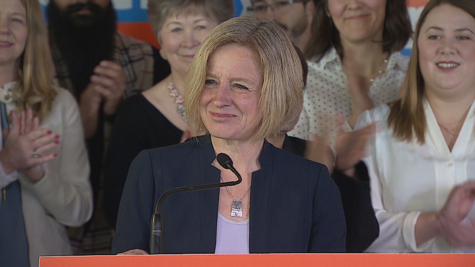 Une femme, tout sourire, avec derrière elle des gens qui applaudissent.