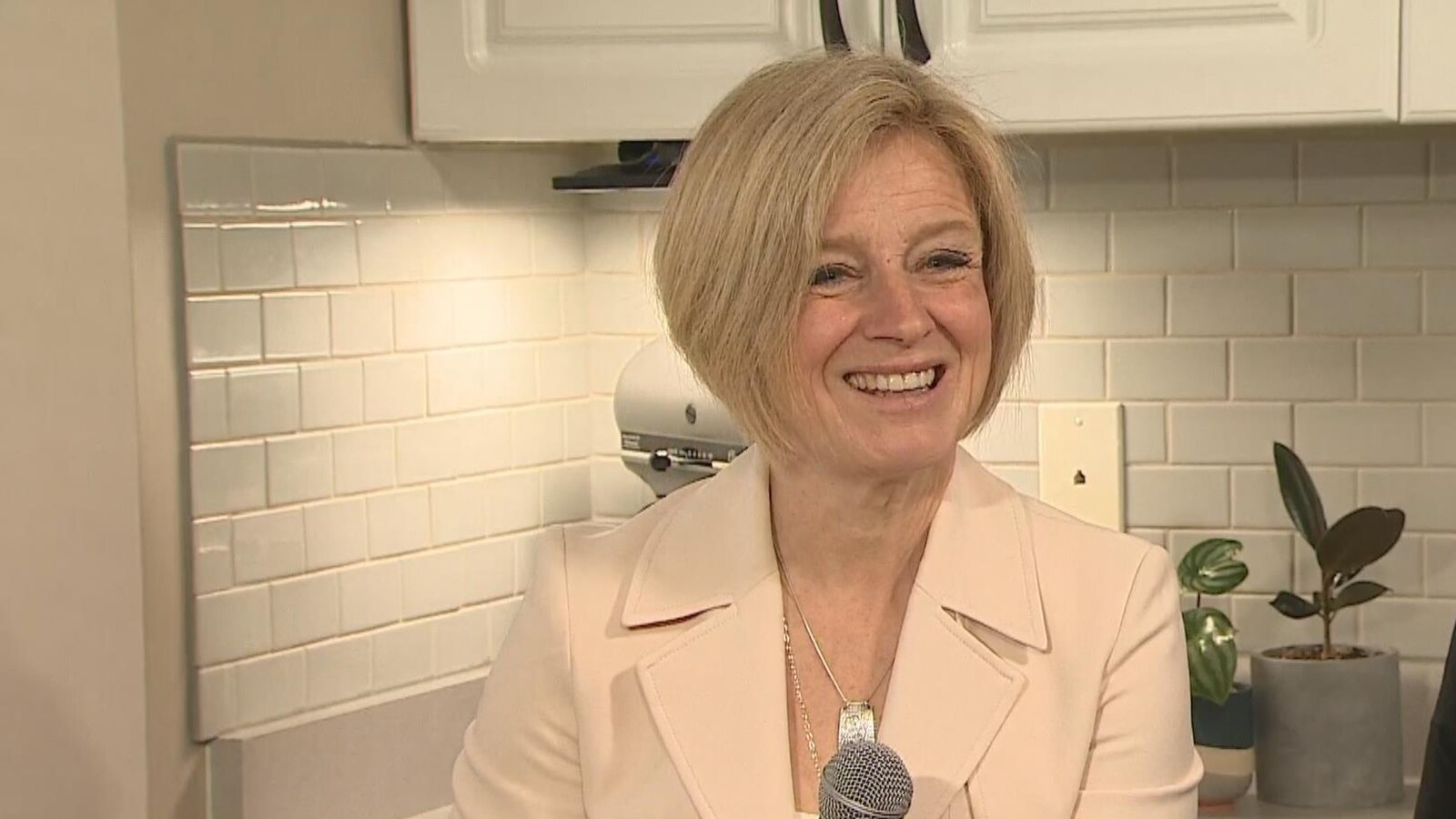 Rachel Notley assise dans une cuisine. Elle tient un micro dans sa main et sourit.