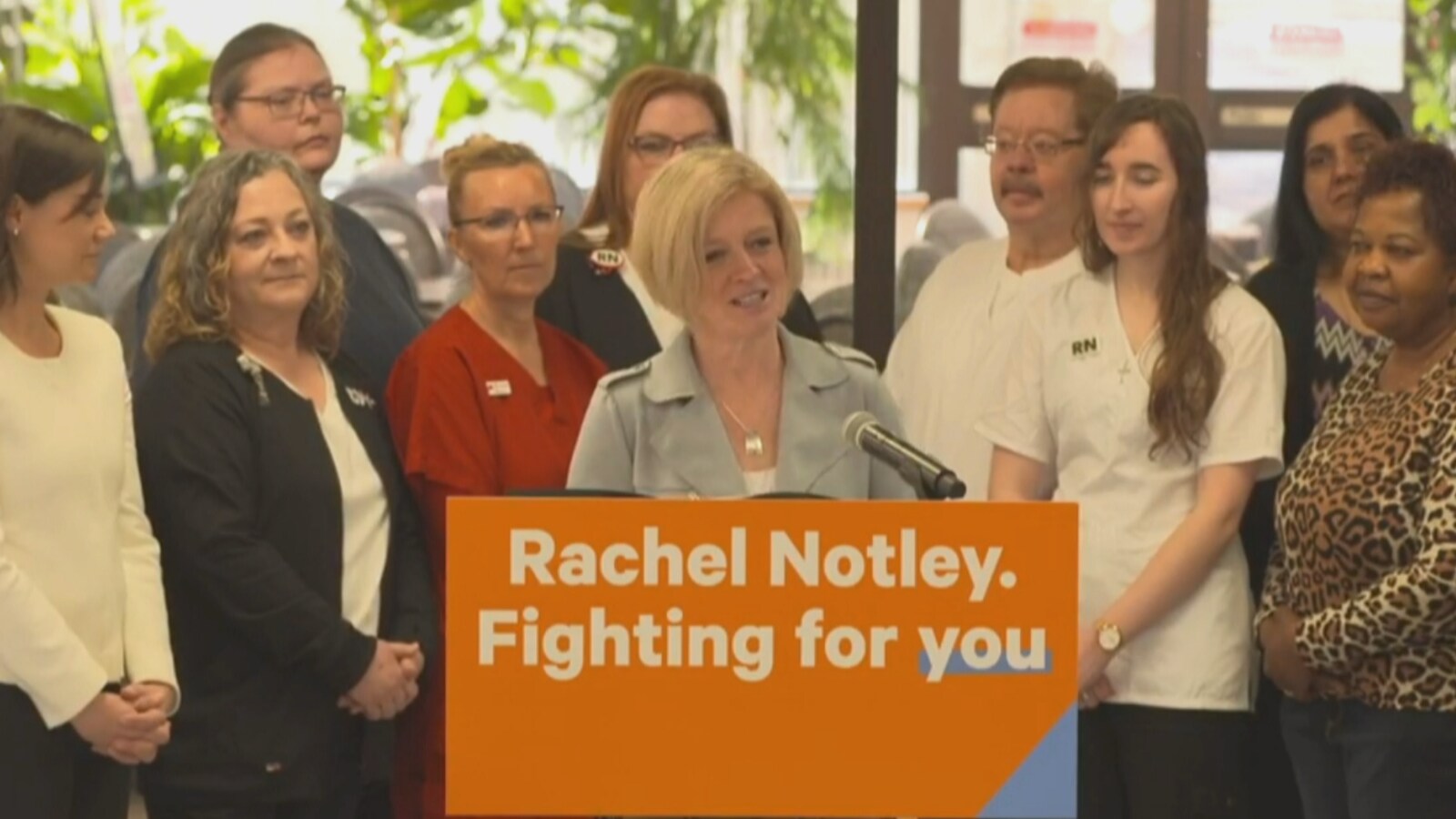Une femme blonde fait un discours derrière un pupitre où il est écrit en anglais: « Rachel Notley. Fighting for you ». Un groupe de personnes se trouvent derrière elle.