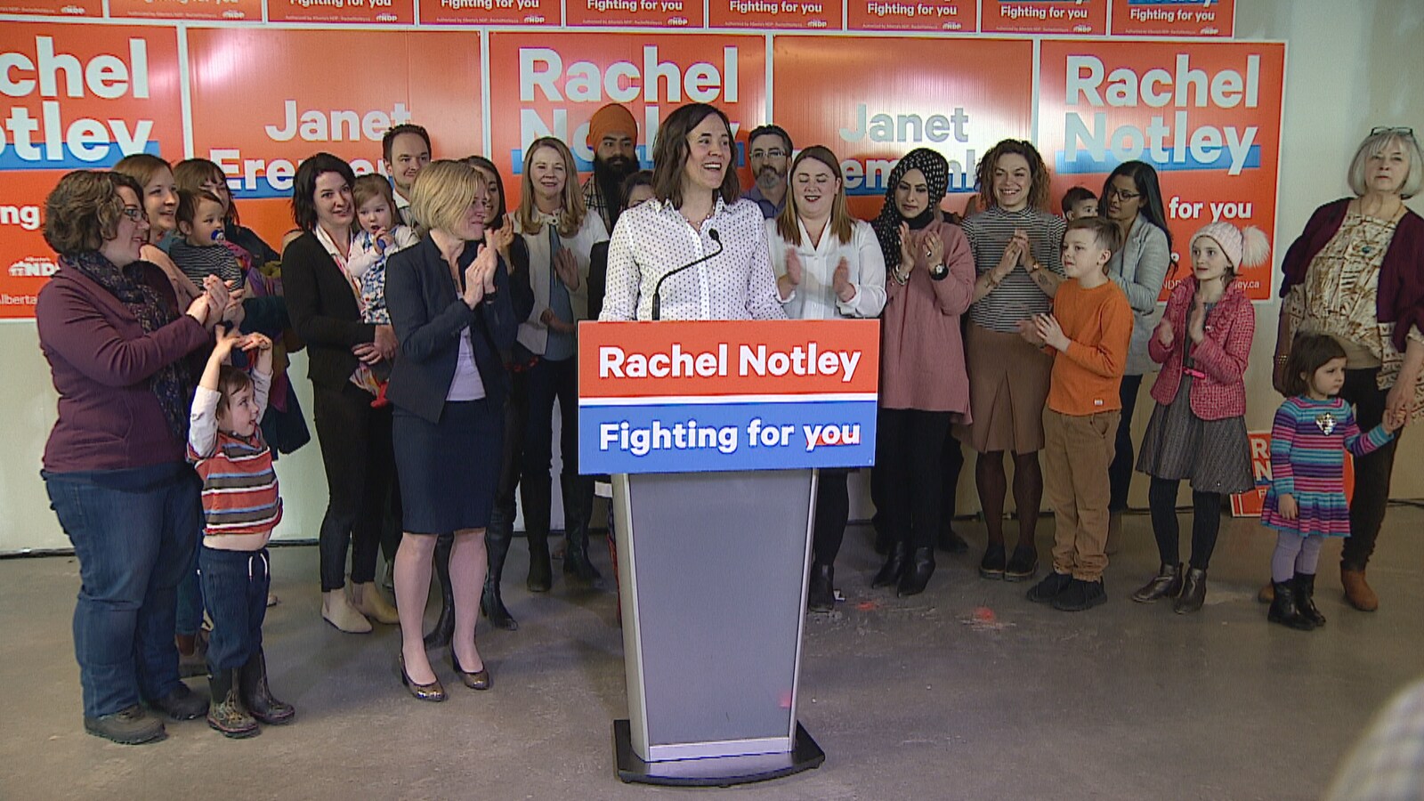 La candidate Janet Eremenko est entourée de Rachel Notley et d'une dizaine de femmes et d'enfants, devant des pancartes électorales.