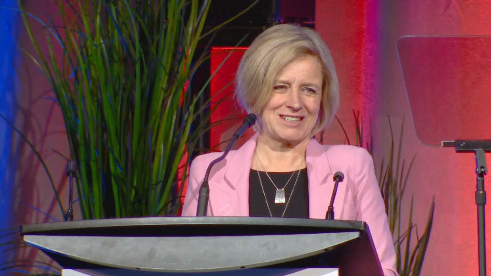 Rachel Notley sourit en parlant à un podium. Elle a les cheveux blonds courts, les yeux clairs, et elle porte un veston rose.