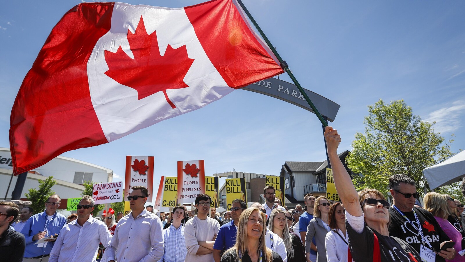 Une femme fait flotter un drapeau canadien. Tout autour d'elle, des personnes tiennent des pancartes en soutien aux pipelines.