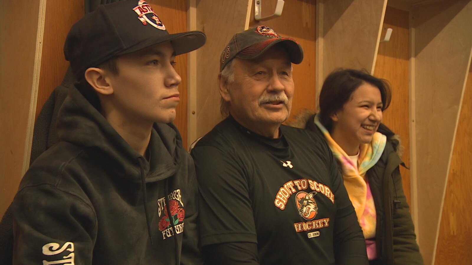 Reggie Leach, au centre : un homme d'un certain âge, portant une casquette et un chandail aux couleurs de son école de hockey "Shoot to Score", entourant de deux jeunes, un garçon et une fille, souriants, dans un vestiaire de hockey.