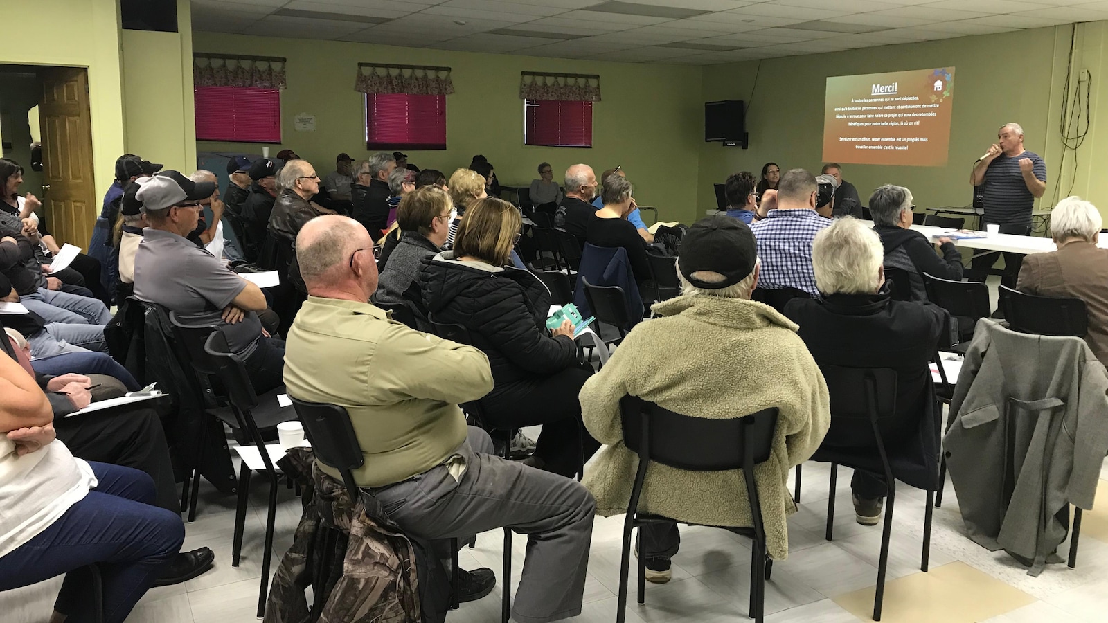 Plusieurs dizaines de personnes écoutent un homme à l'avant de la salle lors d'une présentation.