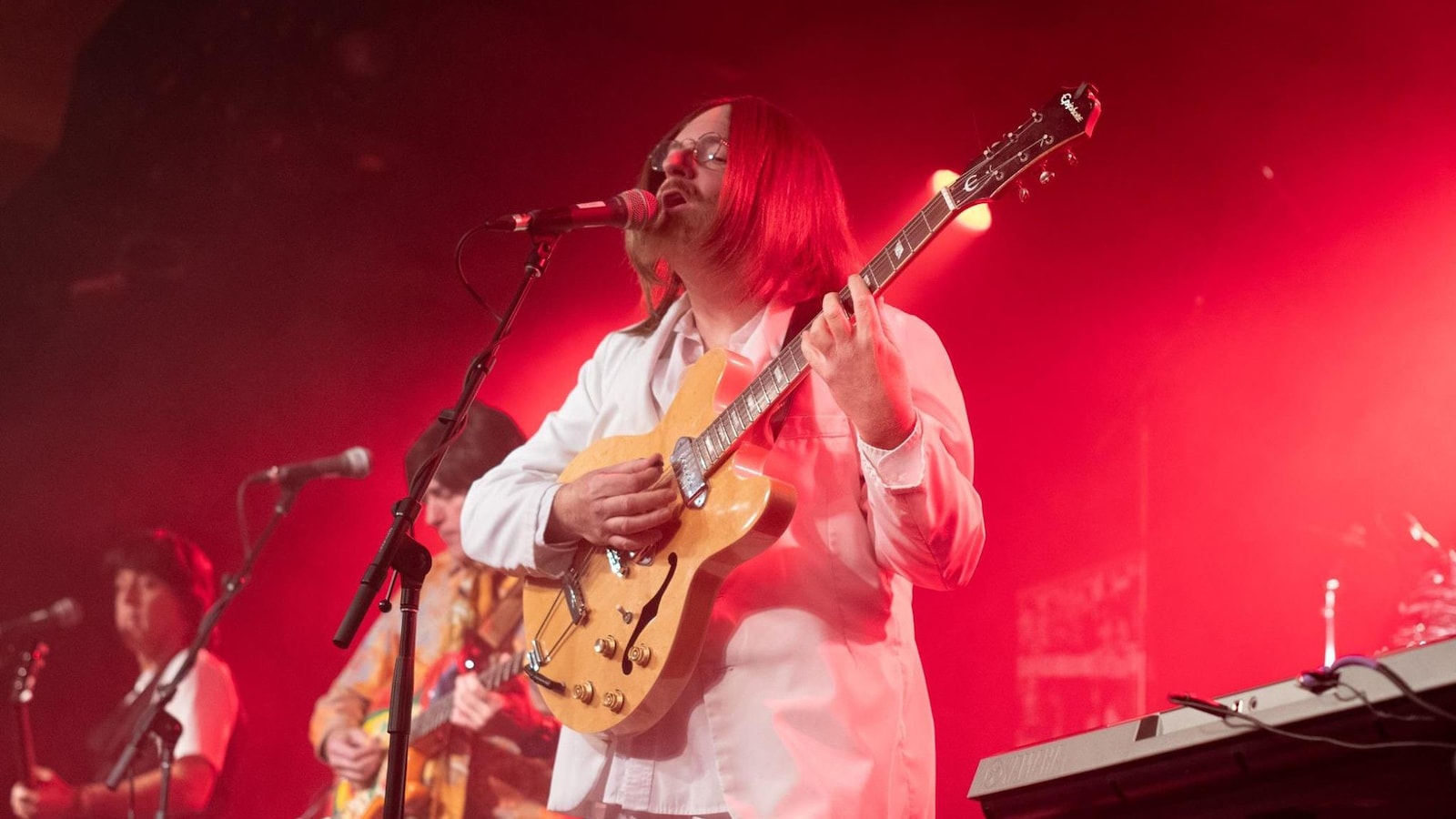 Trois musiciens jouent sur une scène éclairée en rouge. En avant plan, le guitariste chante au micro.