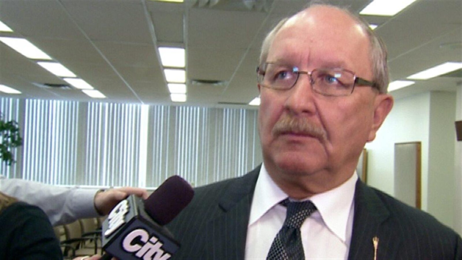 Rick Strankman, a de minces cheveux gris, une moustache et des lunettes. Il est debout et quelqu'un lui tend un micro.