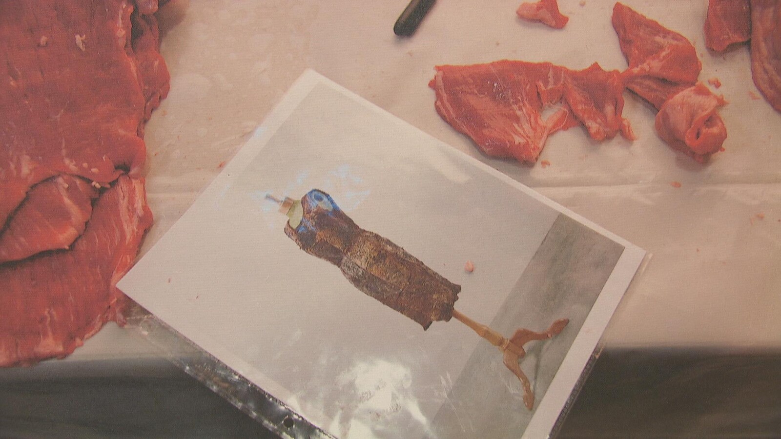 Une photo de robe où on voit une robe en viande séchée est déposée sur une table de boucher. La robe est entourée de morceaux de boeuf. On y voit le manche d'un couteau.
