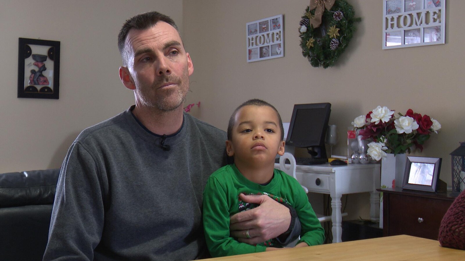 Un homme accorde une entrevue à la caméra derrière une table, un enfant assis sur sa cuisse.