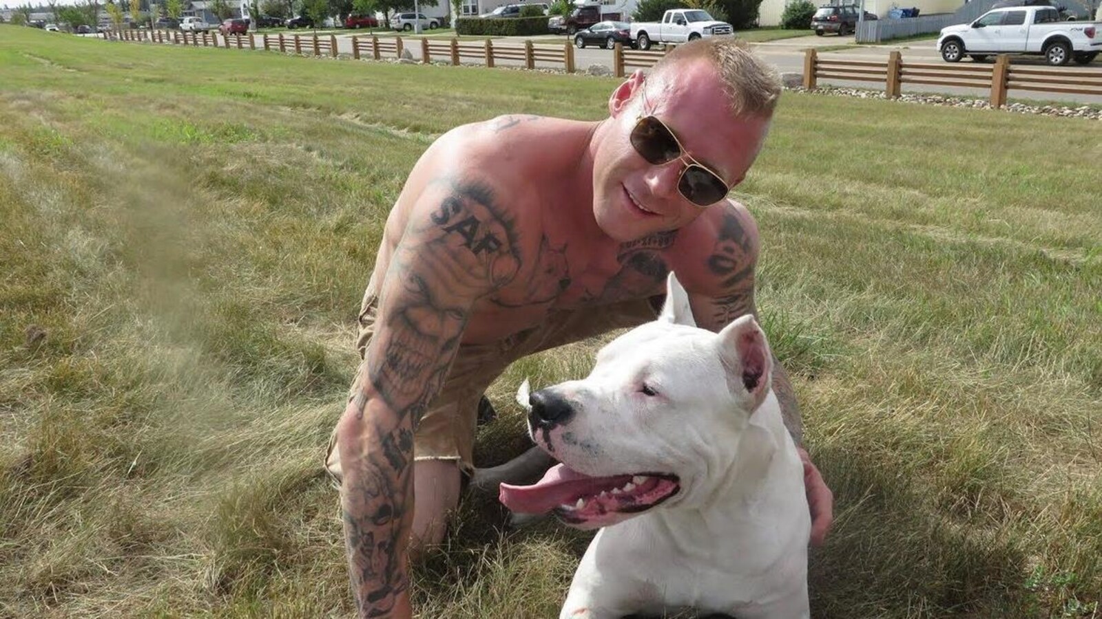 Un homme blond, tatoué, torse nu et portant des lunettes de soleil est accroupi près d'un chien, dans un champ, et regarde la caméra.
