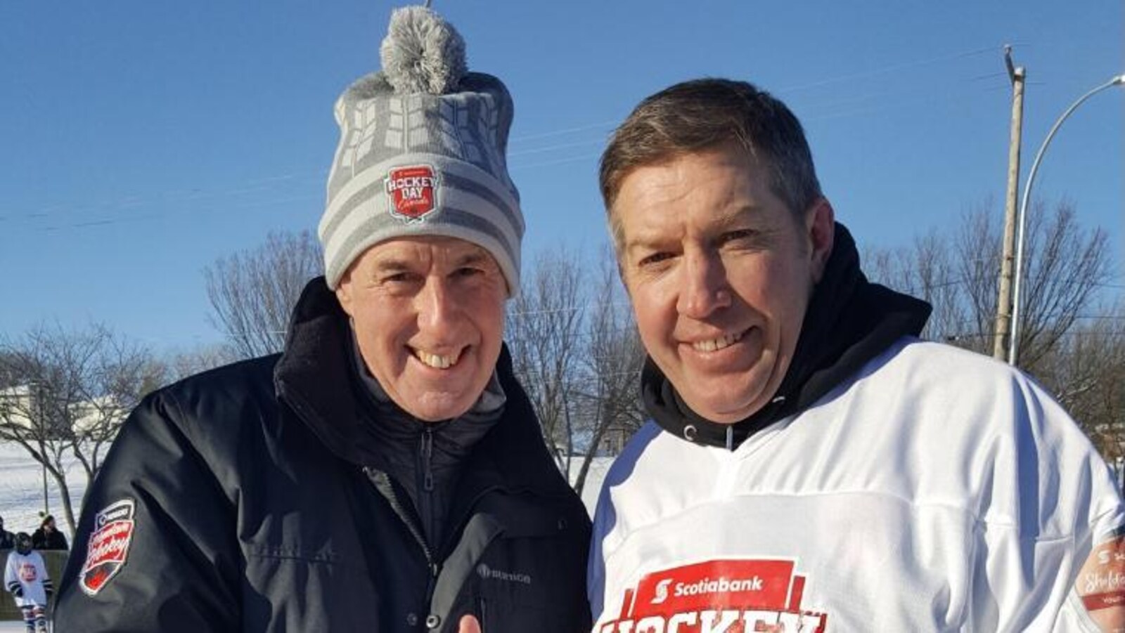 Le commentateur Ron MacLean porte un bonnet et lève le pouce à côté de Sheldon Kennedy qui porte une chemise de hockey.