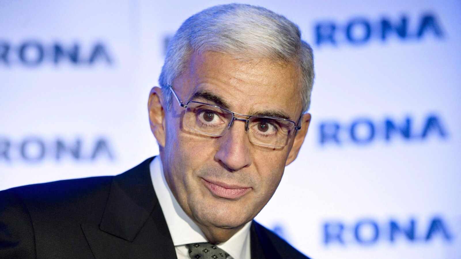 Un homme aux cheveux gris est photographié devant des panneaux sur lesquels on peut lire « Rona ».
