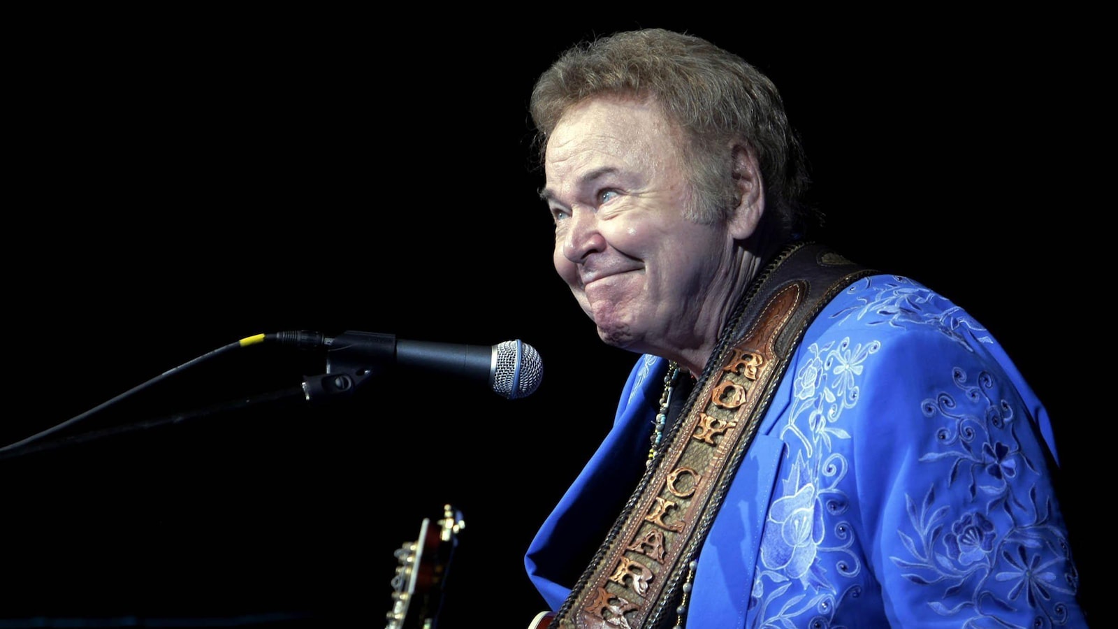 Roy Clark sourit sur scène avec sa guitare.