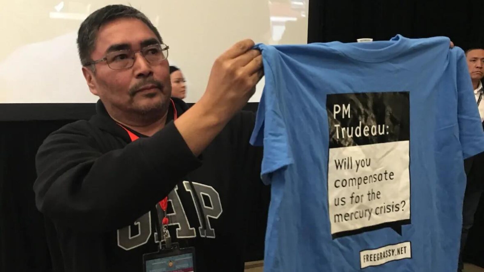 Une homme tenant un chandail sur lequel il est écrit en anglais « Premier ministre Trudeau, allez-vous nous indemniser pour la crise du mercure? »