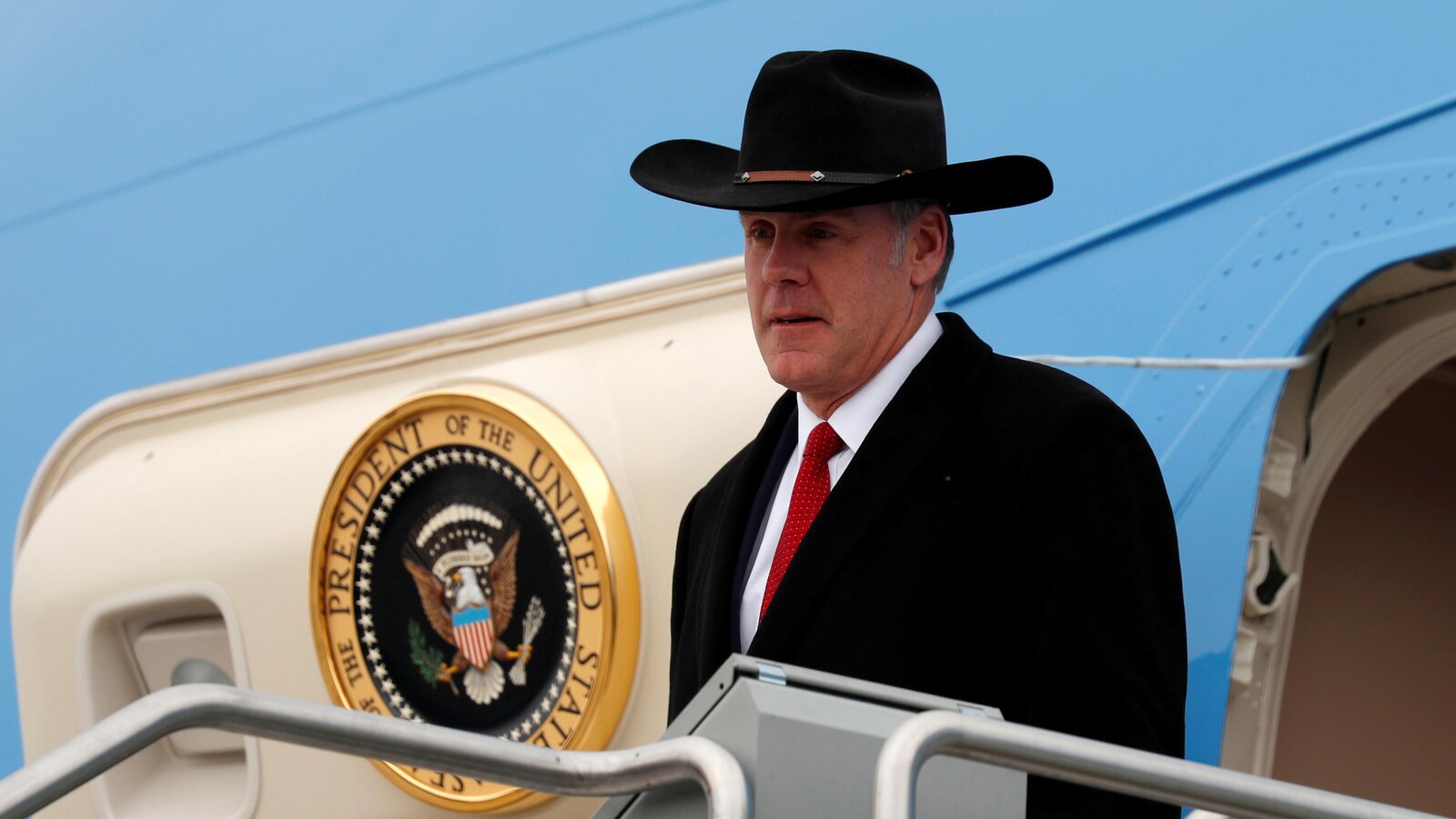 Un homme portant un chapeau de cowboy et un complet avec une cravate sort de avion et s'apprête à descendre un escalier en métal.