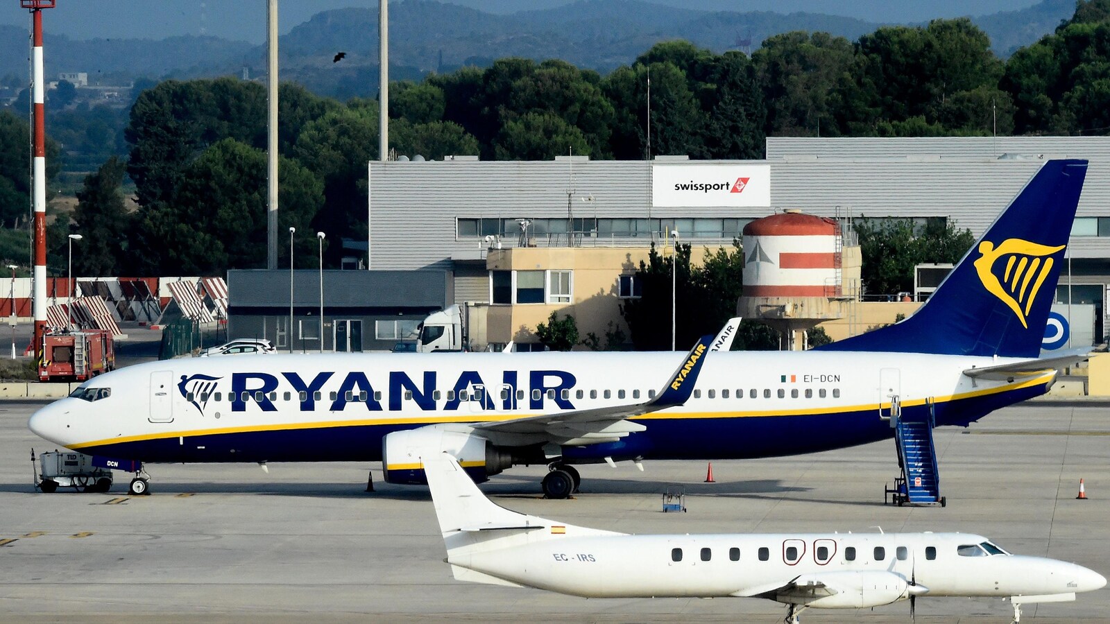 Un avion sur lequel on peut lire « Ryanair » est vu au sol près d'un autre avion. Un escalier permettant aux passagers de quitter l'avion est vu près de l'appareil.