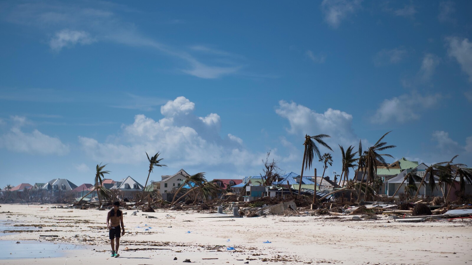 Un homme marche sur la plage lors d'une journée ensoleillée. Autour de lui, les palmiers sont presque déracinés et les habitations sont très endommagées.