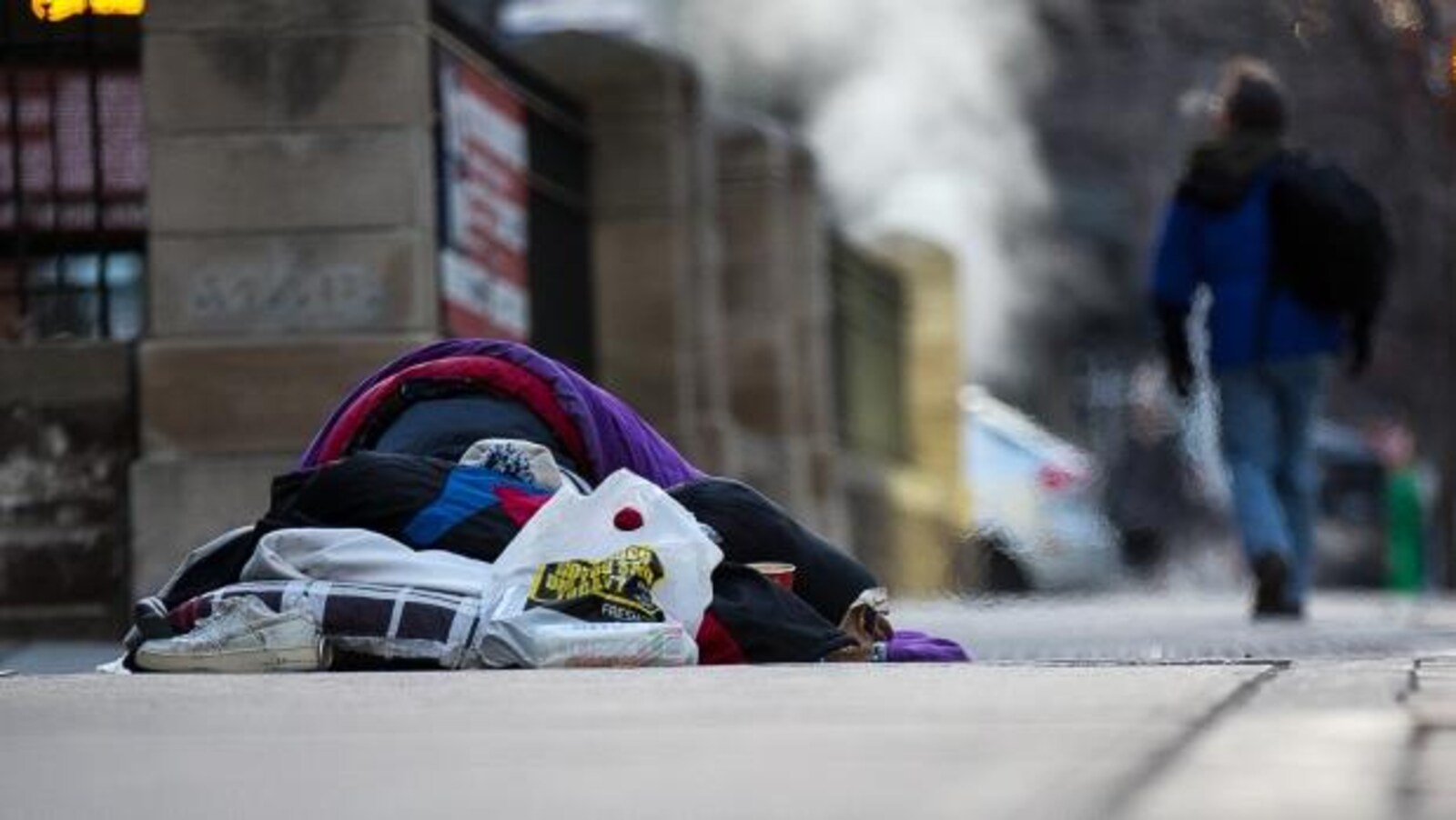 un sans-abri dort dans une rue à même le sol avec un sac de couchage sur lui