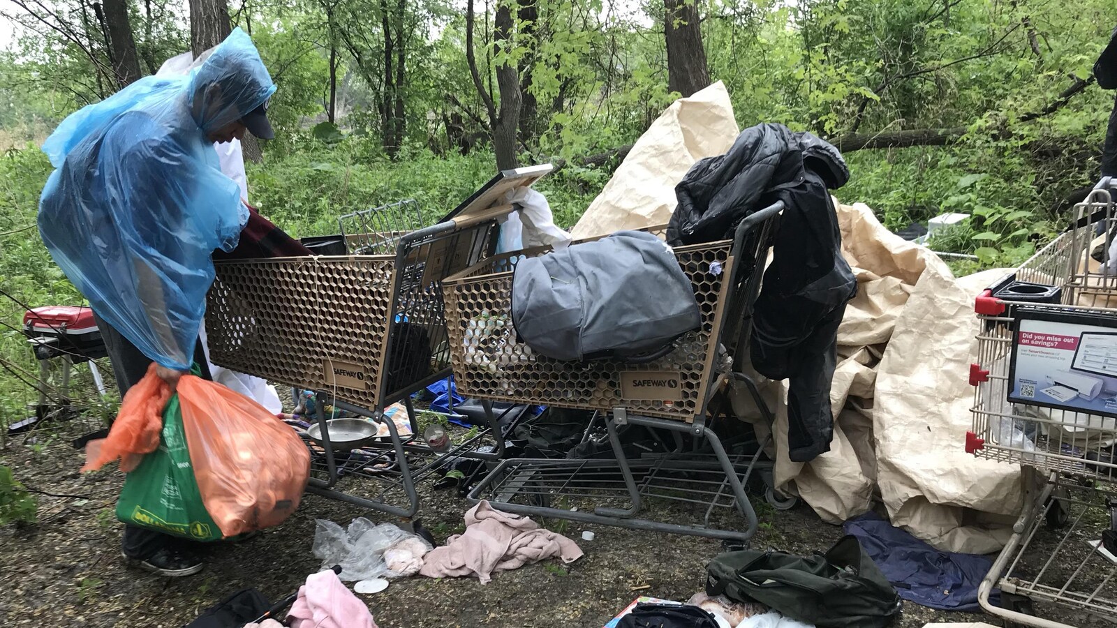 Un homme portant des sacs poubelles remplis de cannettes vides fouille dans des chariots de supermarché contenant de vieux vêtements