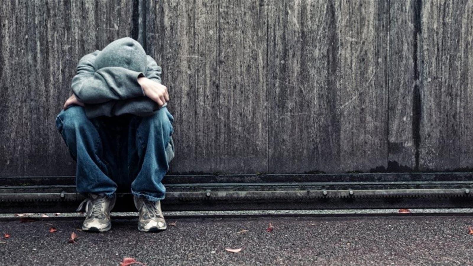 Un jeune adulte recroquevillé sur lui-même, seul devant un grand mur grisâtre.