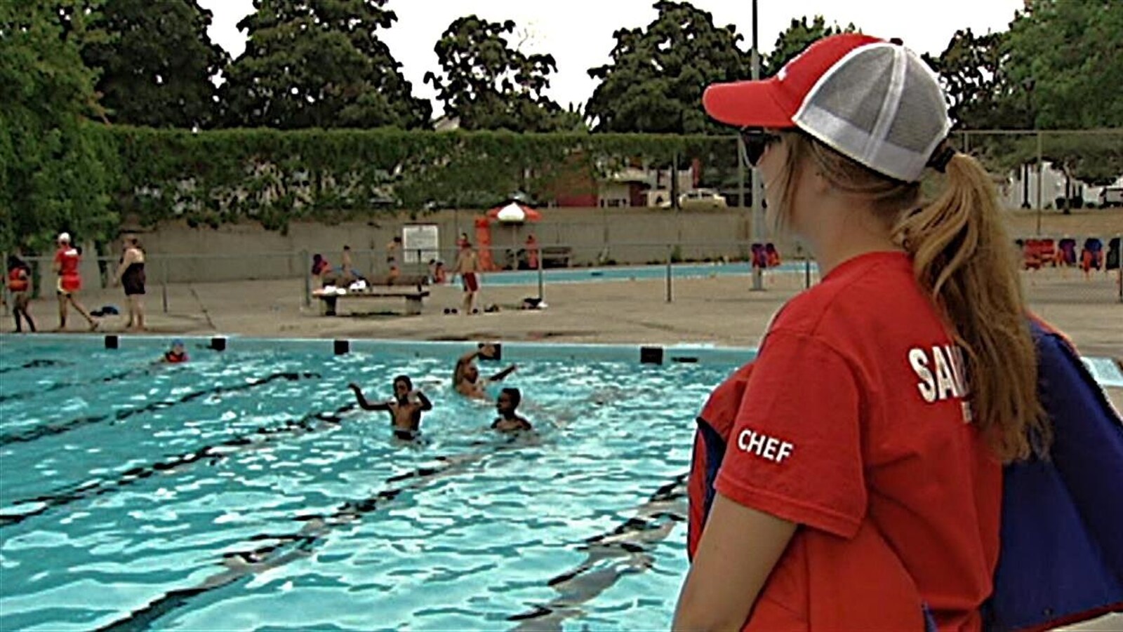 Le jeune femme se tient debout sur le bord de la piscine où se baignent des enfants.