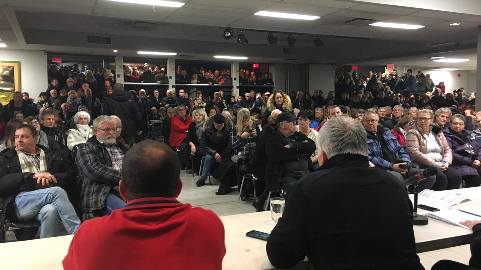 Des centaines de personnes sont assises et debout. En avant, il y a des conseillers municipaux assis à une table.