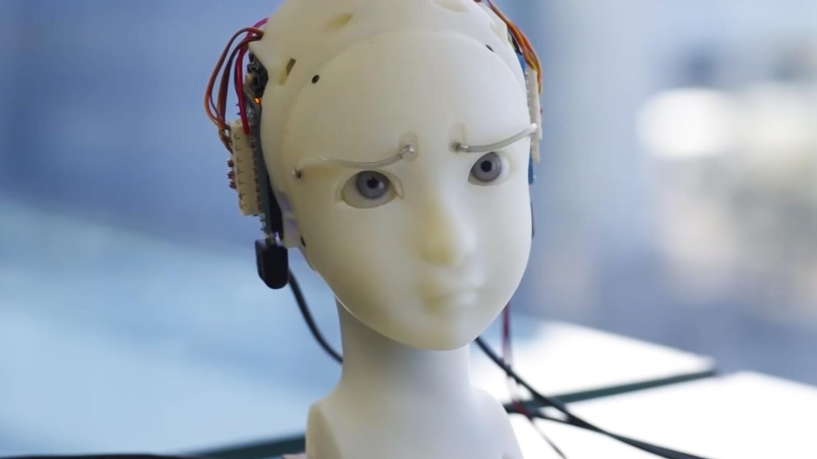 Une capture d'écran montrant un buste blanc aux grands yeux bleus globuleux. Des fils et des pièces électroniques pendant de l'arrière de la tête du buste.