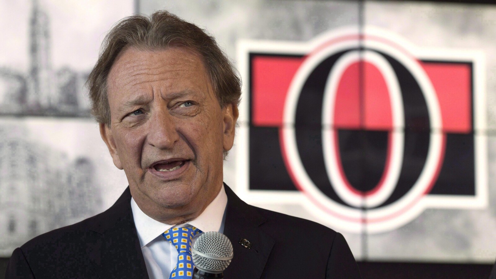 M. Melnyk parle à un micro devant un logo des Sénateurs d'Ottawa.