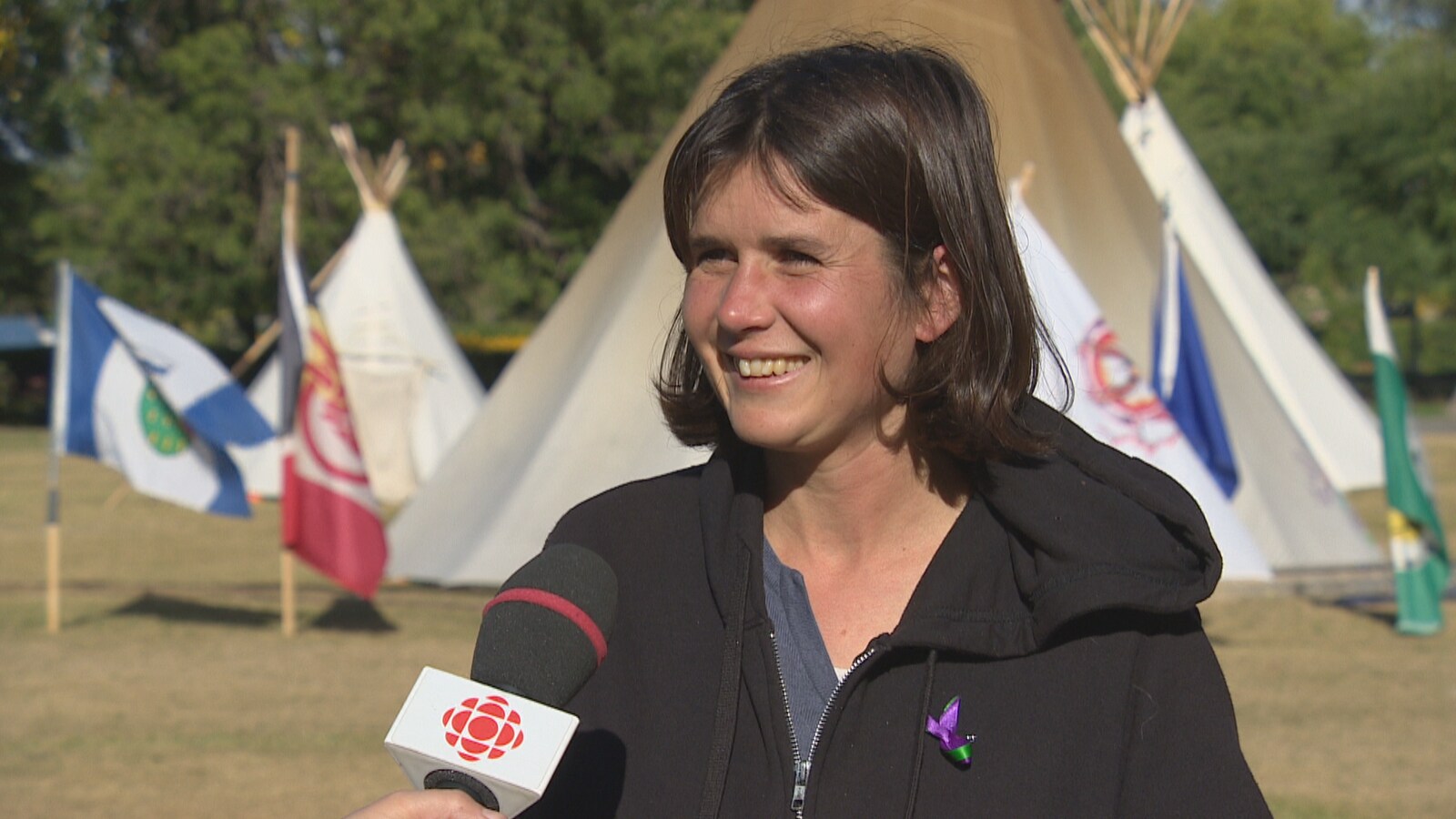 Une jeune femme aux cheveux courts sourit en direction d'une journaliste hors cadre. Derrière elle se trouve le tipi principal, entouré des drapeaux des commaunautés ayant apporté leur soutien au camp.