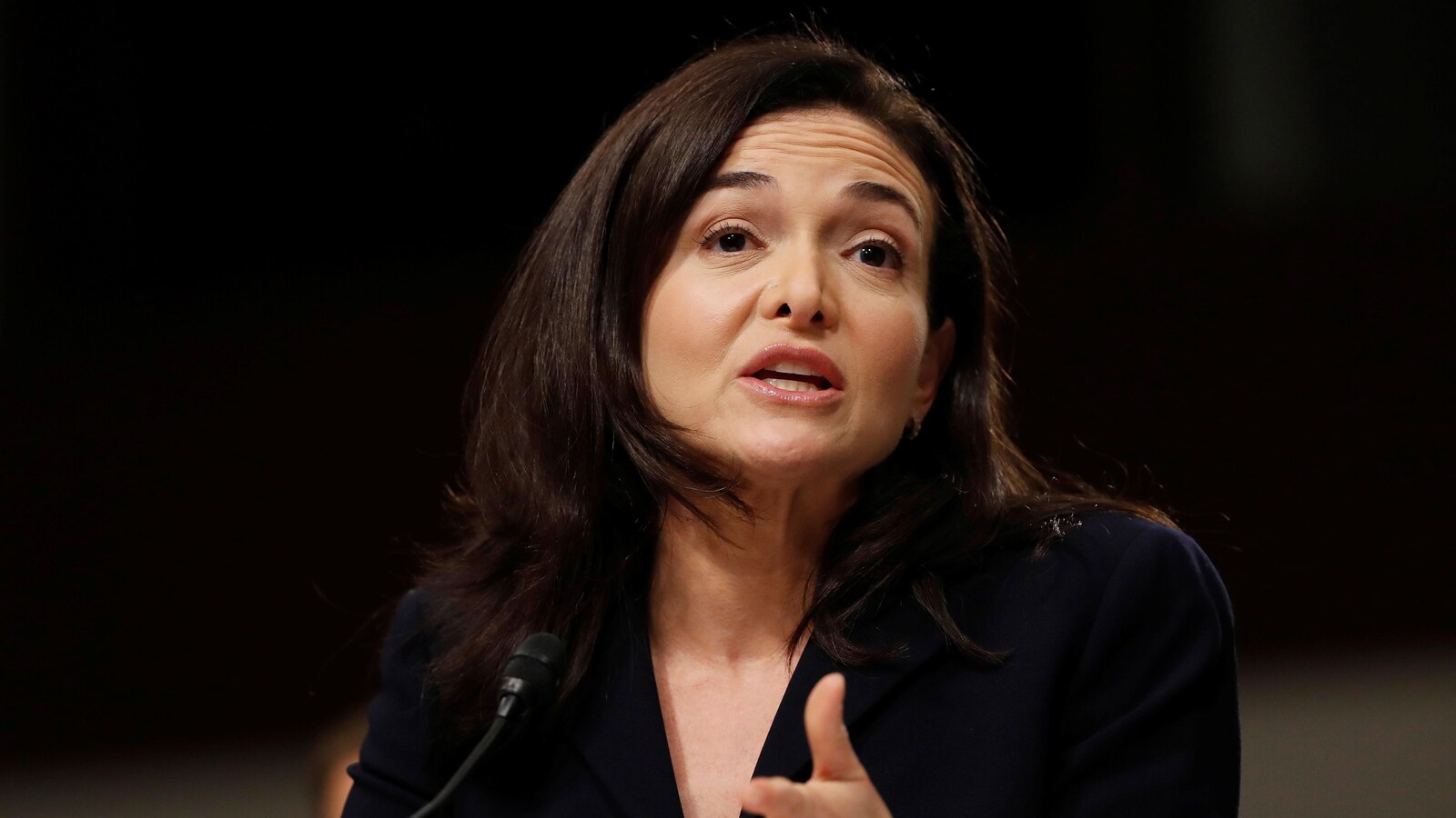 Une photo de Sheryl Sandberg portant un tailleur noir en train de parler devant un micro.
