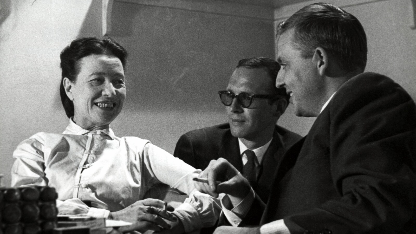 Simone de Beauvoir et Wilfrid Lemoine fumant une cigarette à une petite table. Claude Sylvestre est également assis avec eux, un peu en arrière-plan.