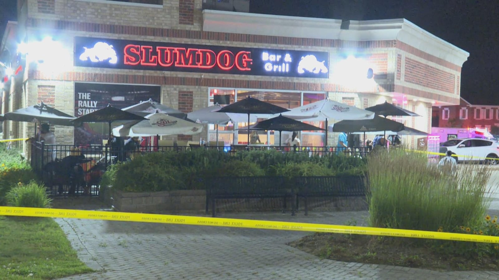 La façade du bar Slumdog où s'est déroulée la fusillade. Il y a un ruban jaune de la police pour ne pas passer, la devanture du bar est éclairée par des néons et il y a une terrasse avec des parasols.