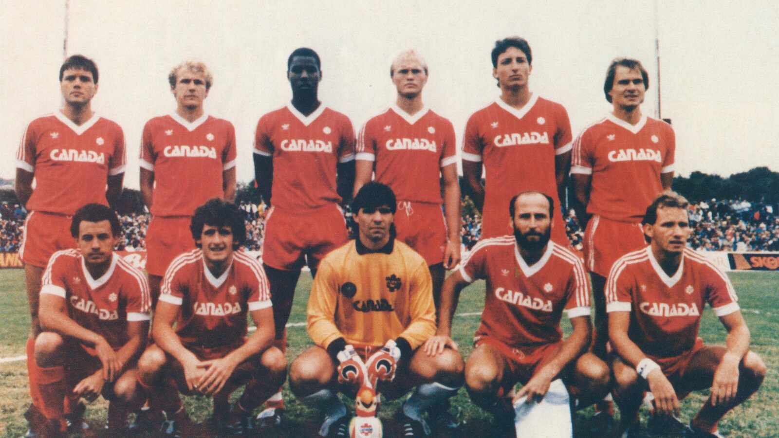 Six joueurs en uniforme sont debout, et cinq sont accroupis devant eux, sur un terrain de soccer.