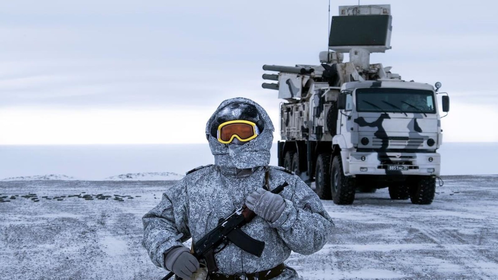 Le soldat est habillé chaudement. On ne voit que ses lunettes de ski dépasser de son capuchon. Il tient un fusil contre lui.