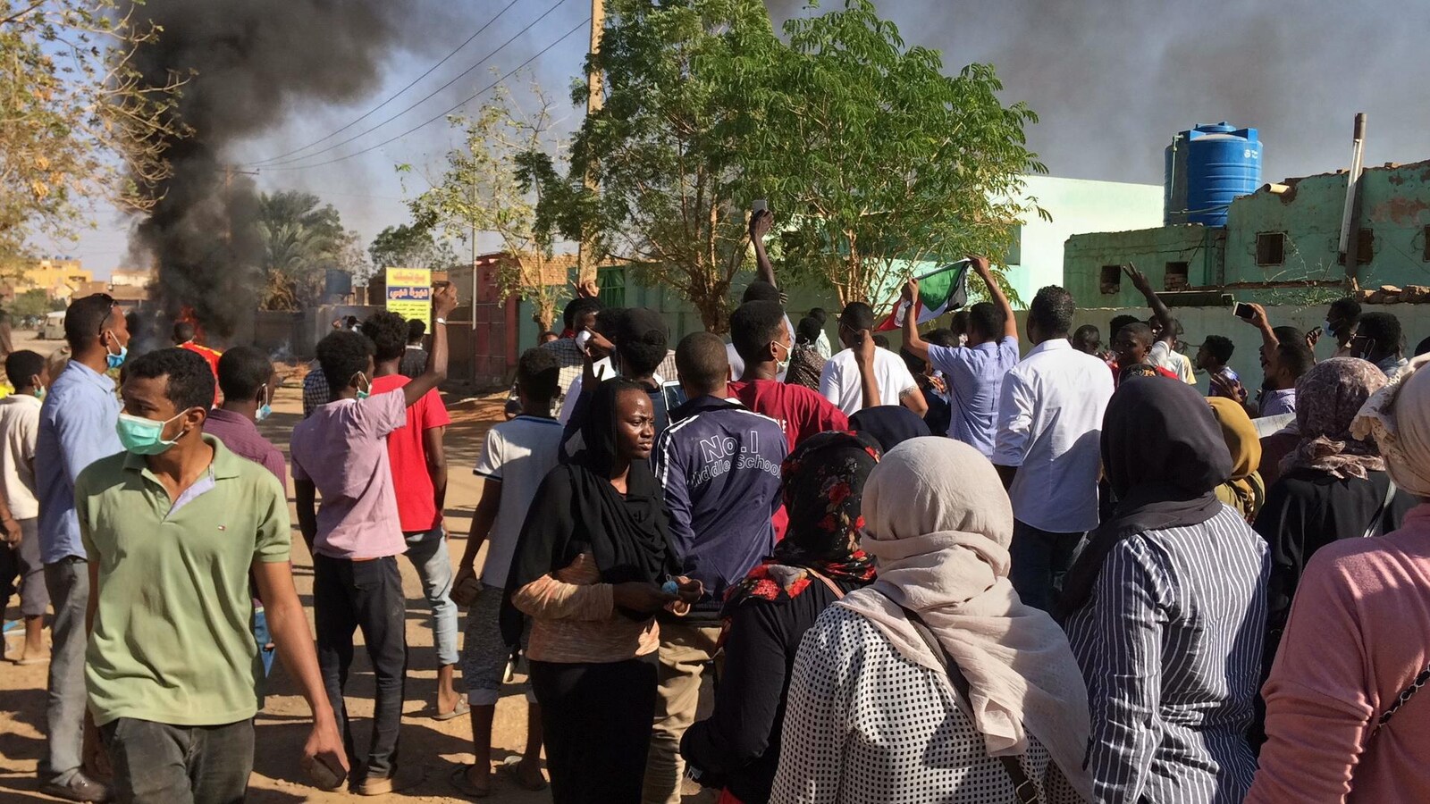 Plusieurs personnes marchent dans une rue de Khartoum. On voit au loin une colonne de fumée noire.