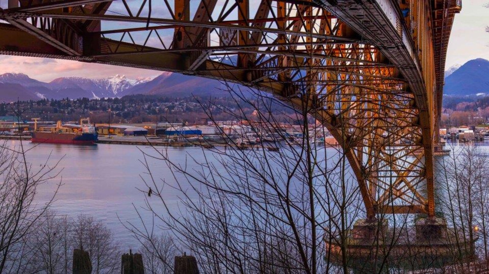 Une photographie des dessous d'un pont métallique avec en arrière-plan la rive nord de Vancouver avec des monts enneigés.