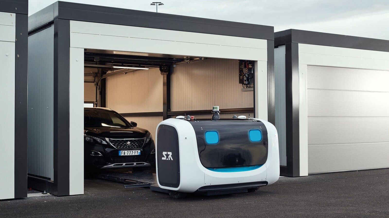 Une photo montrant le robot Stan en train de déposer une voiture dans un garage. Le robot est composé d'une mince plate-forme métallique et d'une grande boîte à l'avant contenant l'équipement électronique.