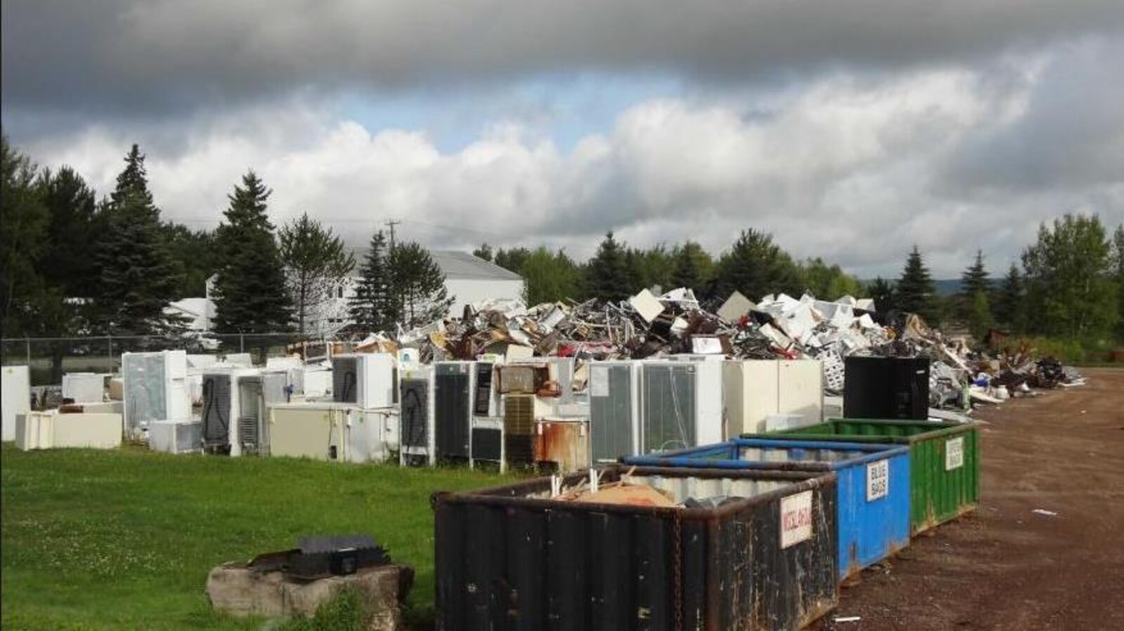 Des conteneurs et des appareils ménagers disposés devant un monticule de déchets sur un terrain clôturé.