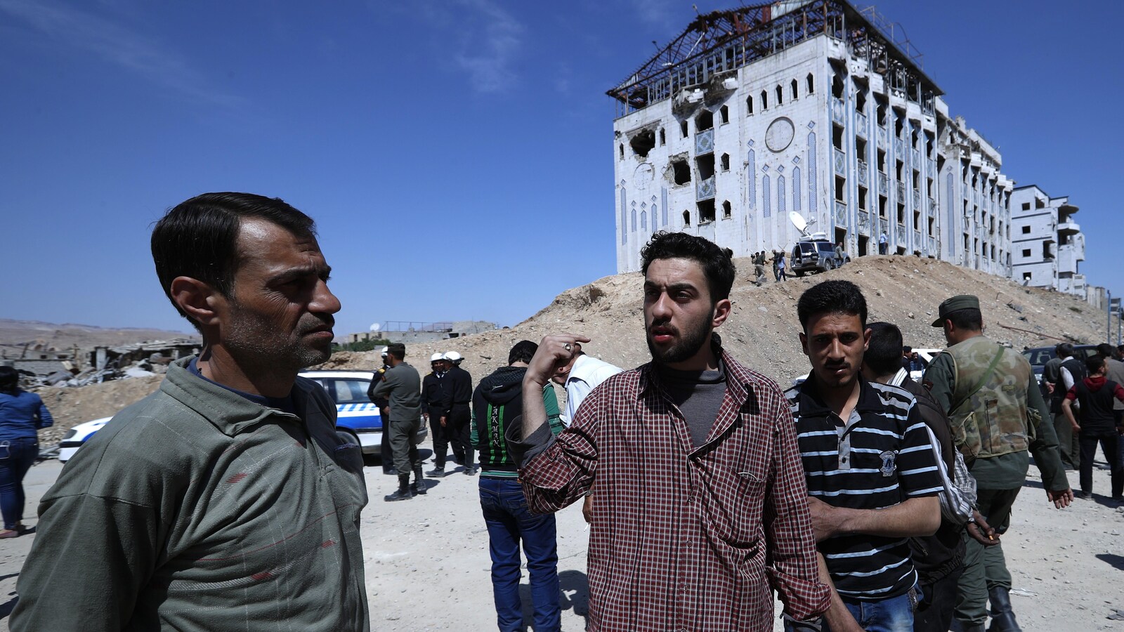 Plusieurs hommes sont rassemblés devant un hôpital de Douma, visiblement endommagé par des obus. Des militaires syriens sont situés tout près.