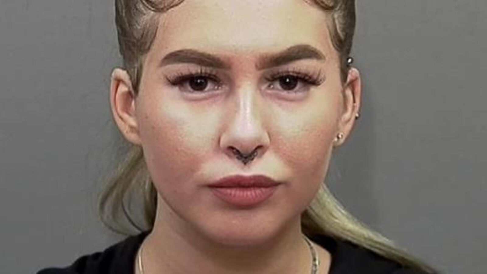 Une jeune fille maquillée, avec des boucles à une oreille et au nez, regarde directement l'objectif de la caméra.