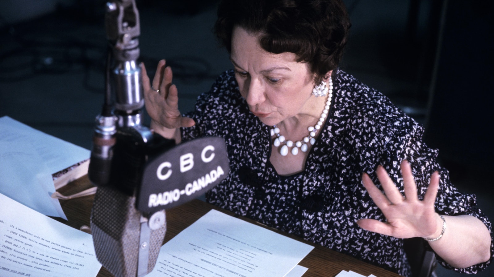 Dans un studio de radio, Lucille Desparois, assise à un pupitre, lit une histoire, les mains en action. Un micro est suspendu devant elle.