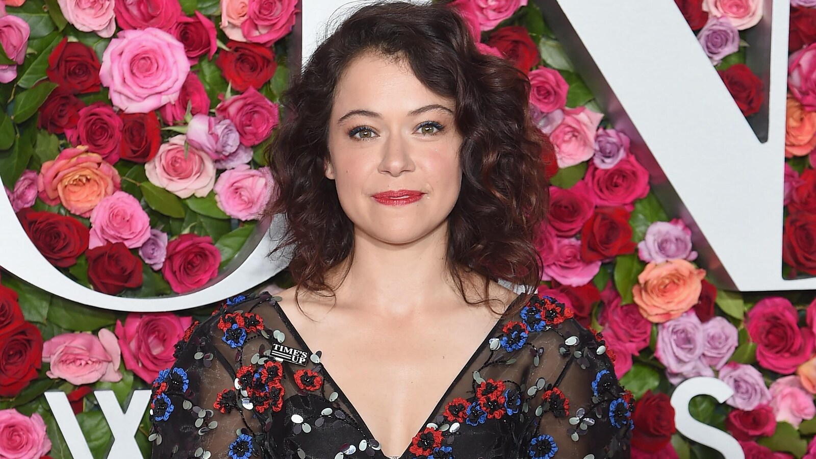 L'actrice canadienne Tatiana Maslany se tient debout devant une mur couvert de roses et sourit.