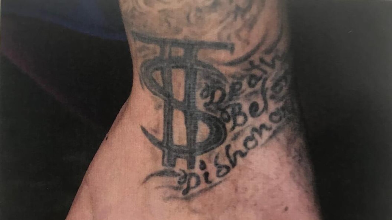 Gros plan sur le dessus d'un poignet gauche sur lequel sont tatouées les lettres «TS» qui débutent un bandeau où l'on peut lire les mots superposés «death», «before» et «dishonor».